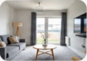 A bright living room with a grey sofa, a round wooden coffee table, a tripod floor lamp and grey curtains at a glass door