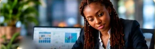 A woman with long braids in a dark blazer working at a laptop showing charts, plants behind her
