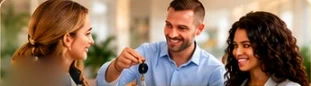 A smiling man in a blue shirt handing a car key to a woman, a second woman beside them