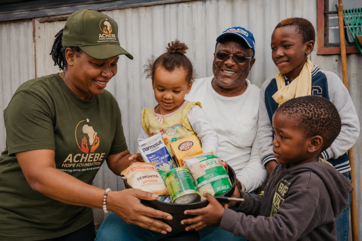 Marie Achebe, in an Achebe Hope Foundation cap and T-shirt, handing a basket of food to a small girl, with her grandfather and two boys beside them