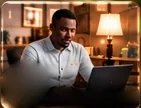 A man in a white shirt writing on a laptop in a lamp-lit room