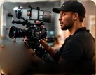 A videographer in a black cap behind a cinema camera rig