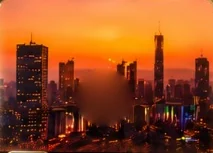 A city skyline at sunset under an orange sky, its lights coming on