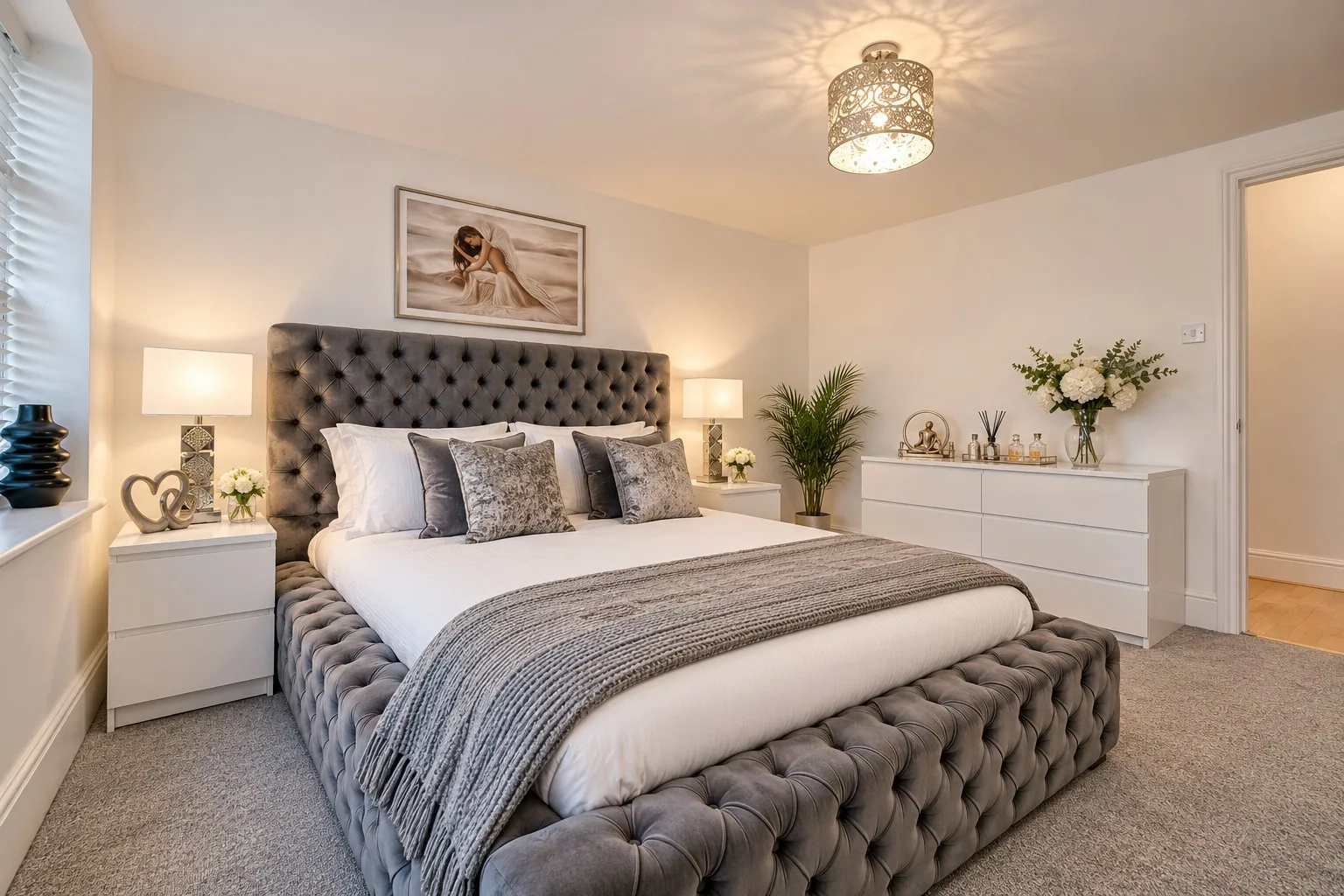 A Marlex bedroom: a grey buttoned bed with a knitted grey throw, white bedside tables and lamps, a painting of a woman above the bed and a pendant light