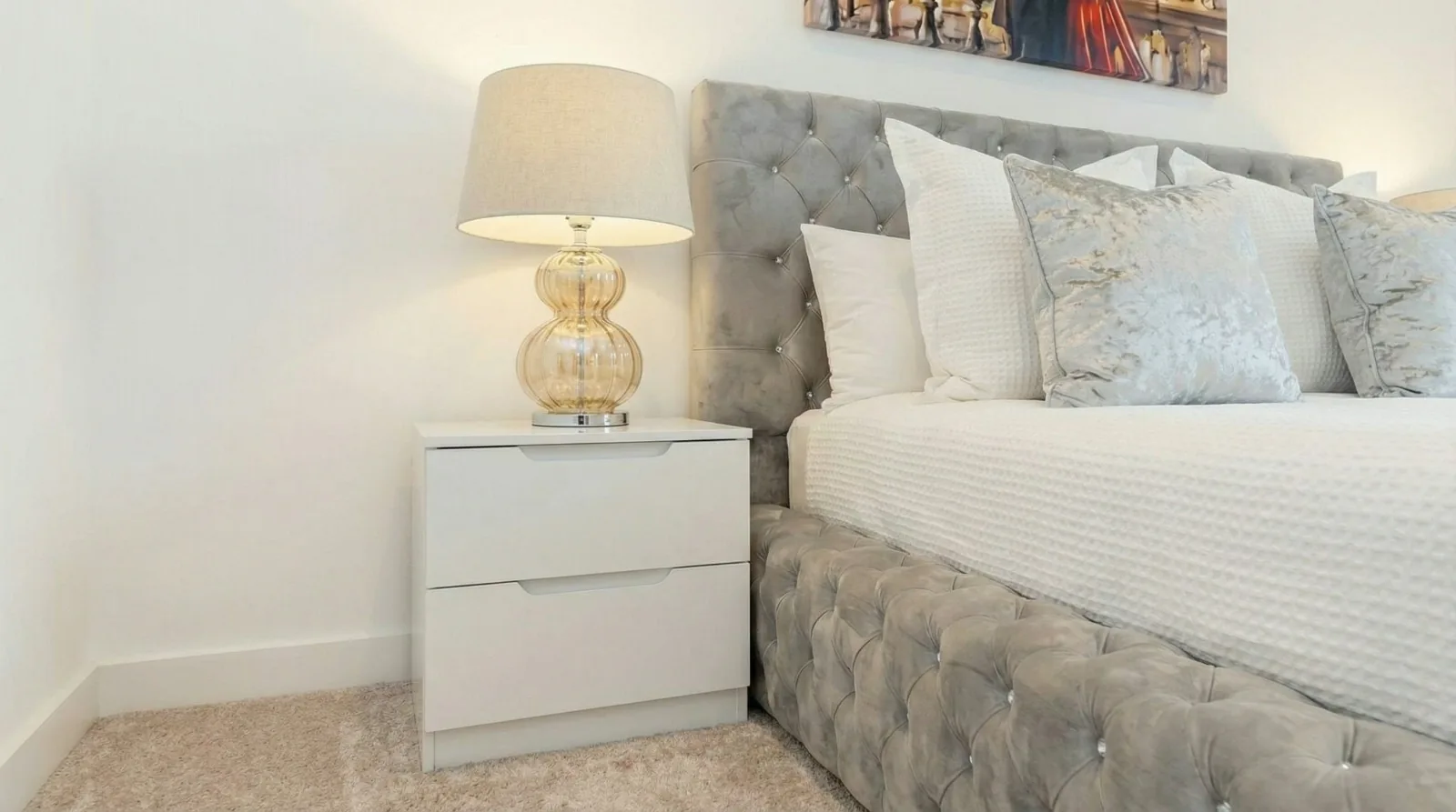 A Marlex apartment bedroom: a gold glass lamp on a white bedside table beside a grey buttoned velvet bed made up in white and silver