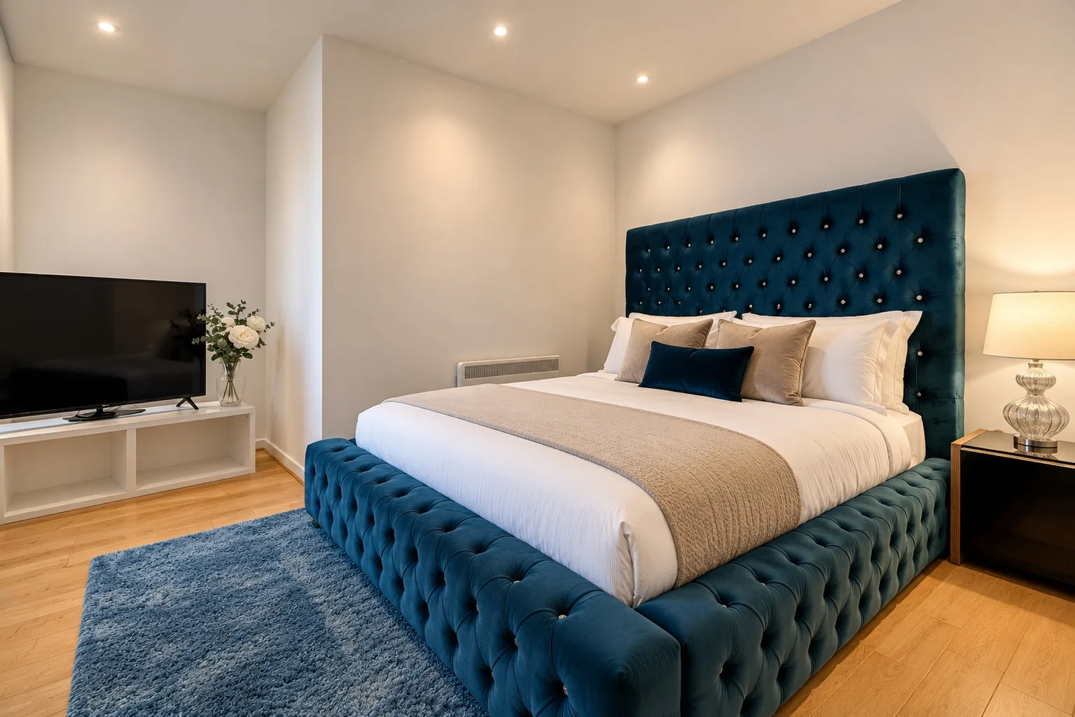 A Marlex bedroom: a deep blue tufted velvet bed made up in white linen, on a blue rug